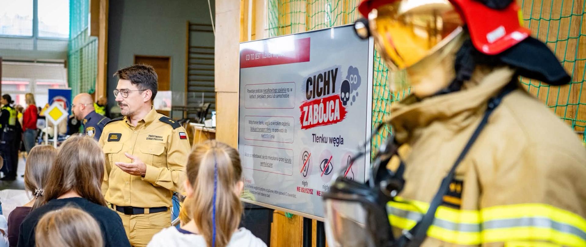 Zdjęcie przedstawia strażaka prowadzącego zajęcia na Akademii Bezpieczeństwa w zakresie zagrożeń ze strony tlenku węgla. Na zdjęciu po prawej widoczny jest manekin strażaka w celu prezentacji ubrania bojowego.