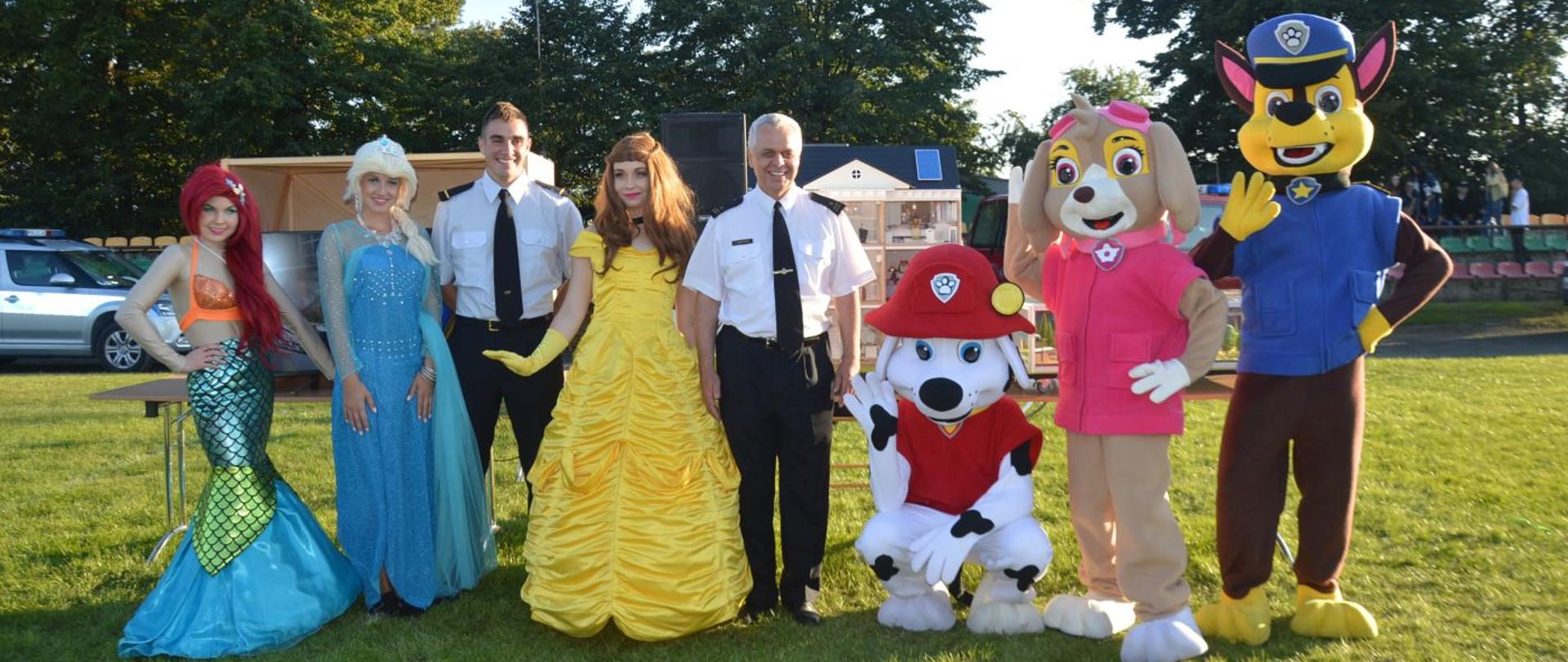Strażacy w krainie bajek. Stadion sportowy w Jutrosinie. Na pierwszym planie strażacy z Komendy Powiatowej PSP w Rawiczu w towarzystwie postaci z bajek, m.in. bohaterów "Psiego patrolu". Za nimi symulator zagrożeń, drzewa i trybuny.
