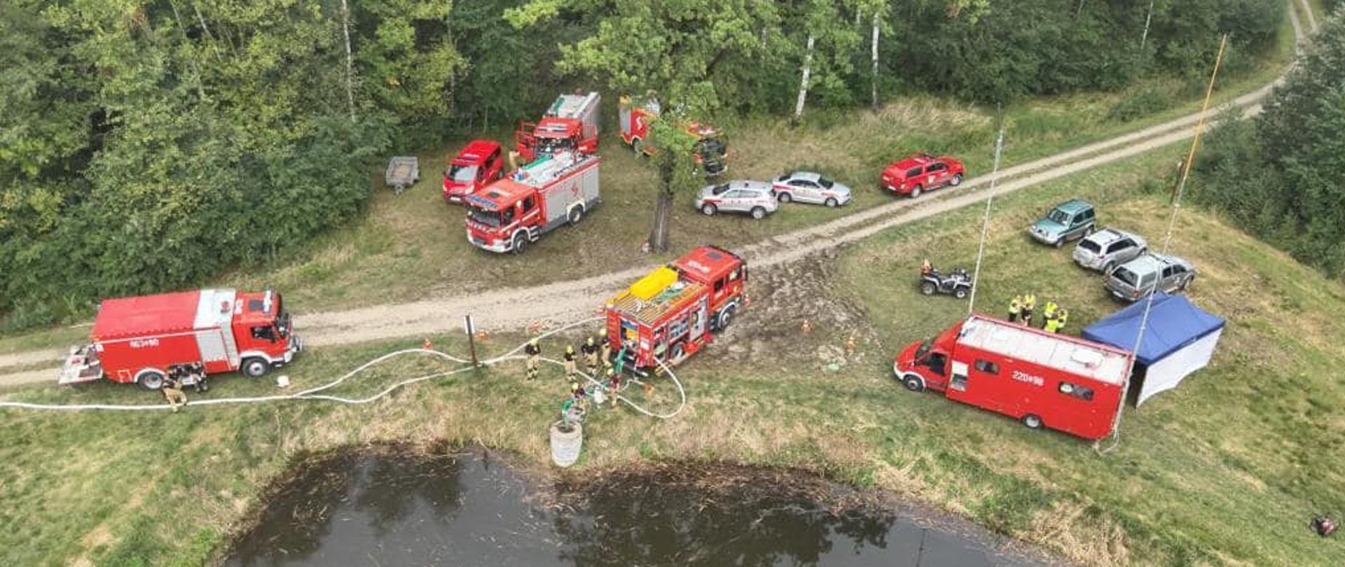 Na zdjęciu widać z "lotu ptaka" 8 samochodów strażackich, 5 cywilnych, kład oraz namiot i kilku strażaków na tle lasu i zbiornika wodnego