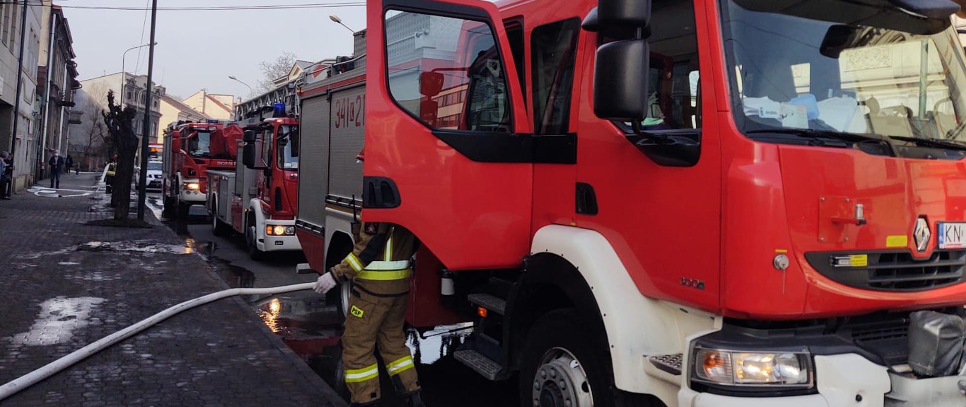 Na ulicy Długosza stoją samochody pożarnicze uczestniczące w zdarzeniu.Z jednego z nich rozwinięta jest linia wężowa. Obok samochodu stoi strażak. Drzwi samochodu są otwarte.