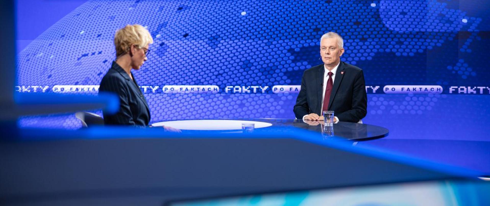 Zdjęcie przedstawia wnętrze studia telewizyjnego. Minister Tomasz Siemoniak (po prawej) oraz redaktor Anita Werner (po lewej) siedzą przy stole. Na stole stoją szklanki z wodą. Za ministrem paski z nazwą programu „Fakty po Faktach” oraz niebieskie tło z konturami Europy. Na dole i na górze zdjęcia widać fragmenty monitorów telewizyjnych.