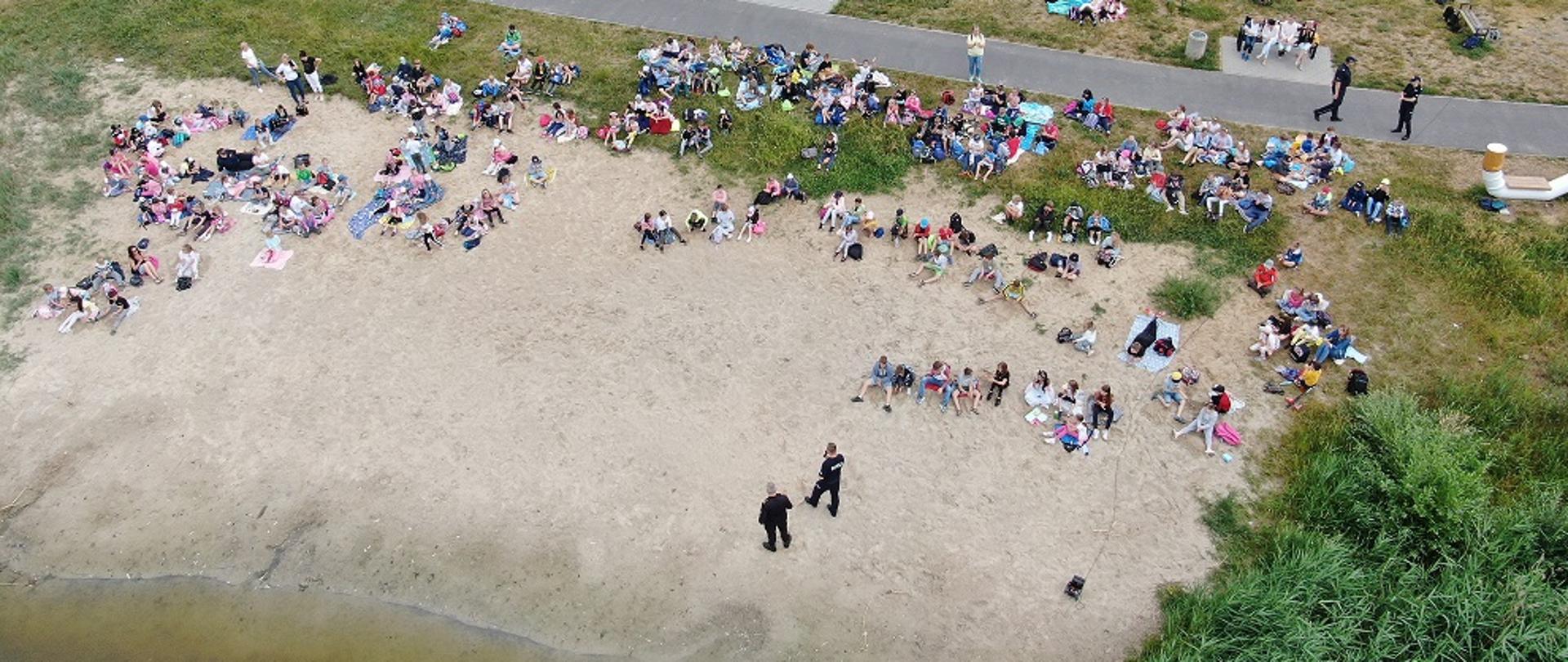 Zdjęcie przedstawia ujęcie z drona nad plażą. Na zdjęciu znajdują się funkcjonariusze oraz dzieci.