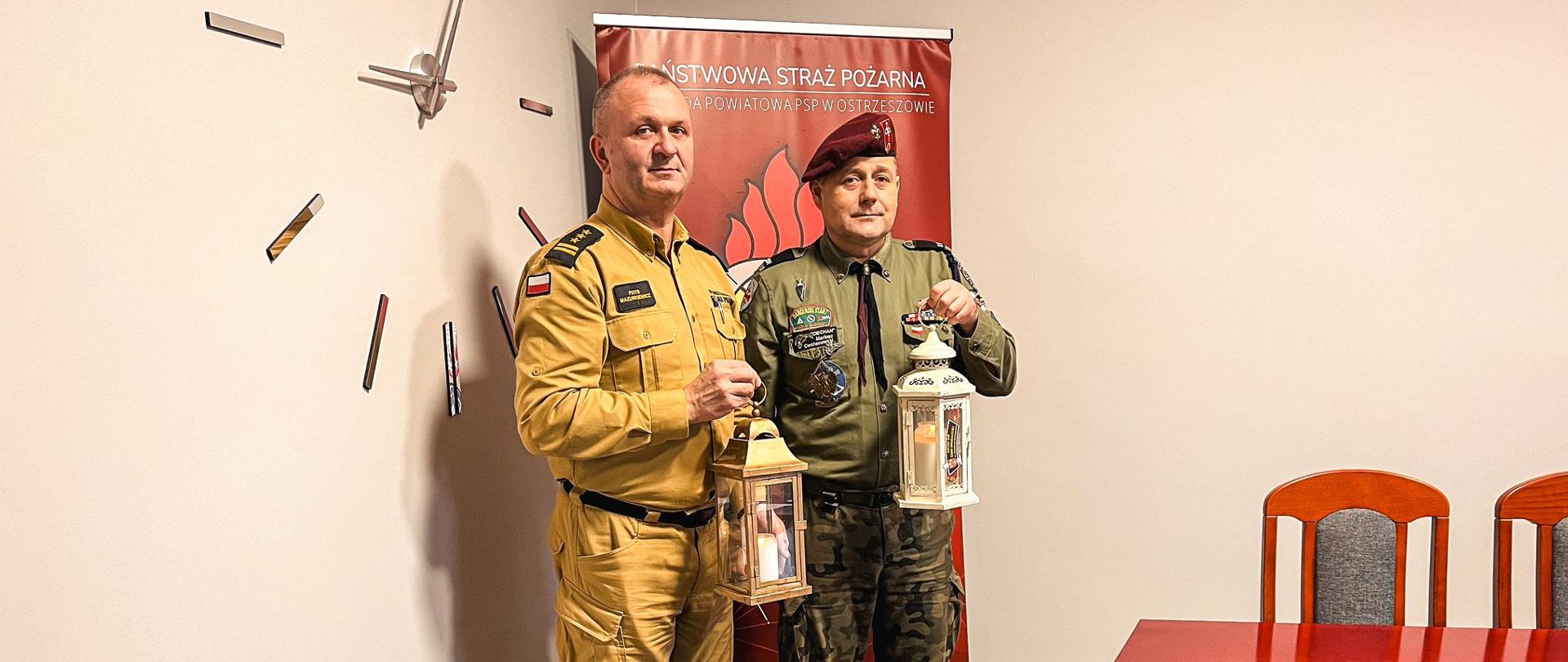 Komendant Powiatowy odebrał od Komendanta Harcerzy symboliczne Światełko Pokoju poprzez zapalenie lampionu. Zdjęcie wykonane w budynku komendy. Za osobami baner Komendy Powiatowej Państwowej Straży Pożarnej w Ostrzeszowie.
