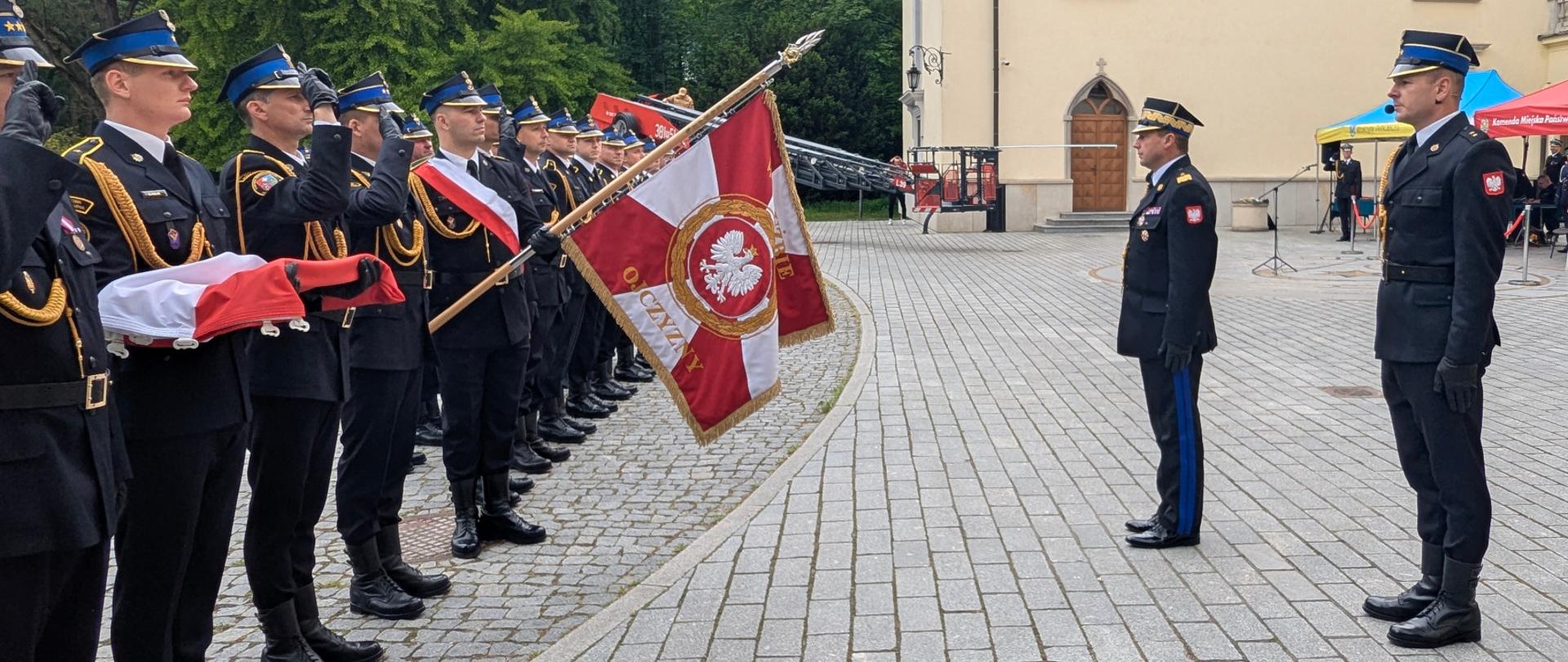 Szpaler strażaków w galowych mundurach stoi na placu, z polską flagą niesioną przez dwóch z nich. Po prawej stronie widać dwóch strażaków stojących naprzeciwko siebie,. Strażak po prawej stronie trzyma pochylony sztandar. W tle rozstawiony jest czerwony namiot i gromadzą się inni ludzie.