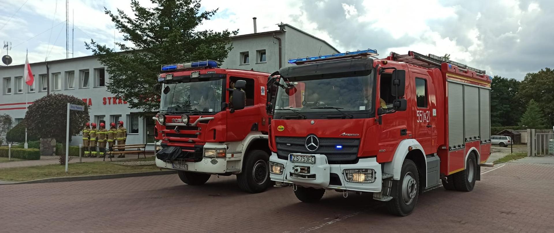 Zdjęcie przedstawia dwa samochody ratowniczo-gaśnicze z włączonymi światłami mijania oraz sygnałami świetlnymi. auta znajduja się przed budynkiem komendy PSP w Świdwinie.