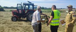 Zdjęcie przedstawia scenę na skoszonym, trawiastym polu w słoneczny dzień. W tle, po lewej stronie, stoi czerwony ciągnik rolniczy marki Ursus z podpiętą maszyną rolniczą. Na pierwszym planie widoczne są cztery osoby prowadzące rozmowę: Starszy mężczyzna w jasnej koszuli z krótkim rękawem i szarych spodniach, zwrócony tyłem do obiektywu. Mężczyzna w kamizelce odblaskowej z zielonym logo instytucji na plecach. Mężczyzna w beżowym mundurze i czapce z daszkiem, trzymający w ręku ulotki lub broszury. Czwarta osoba, częściowo zasłonięta, stojąca bliżej ciągnika.