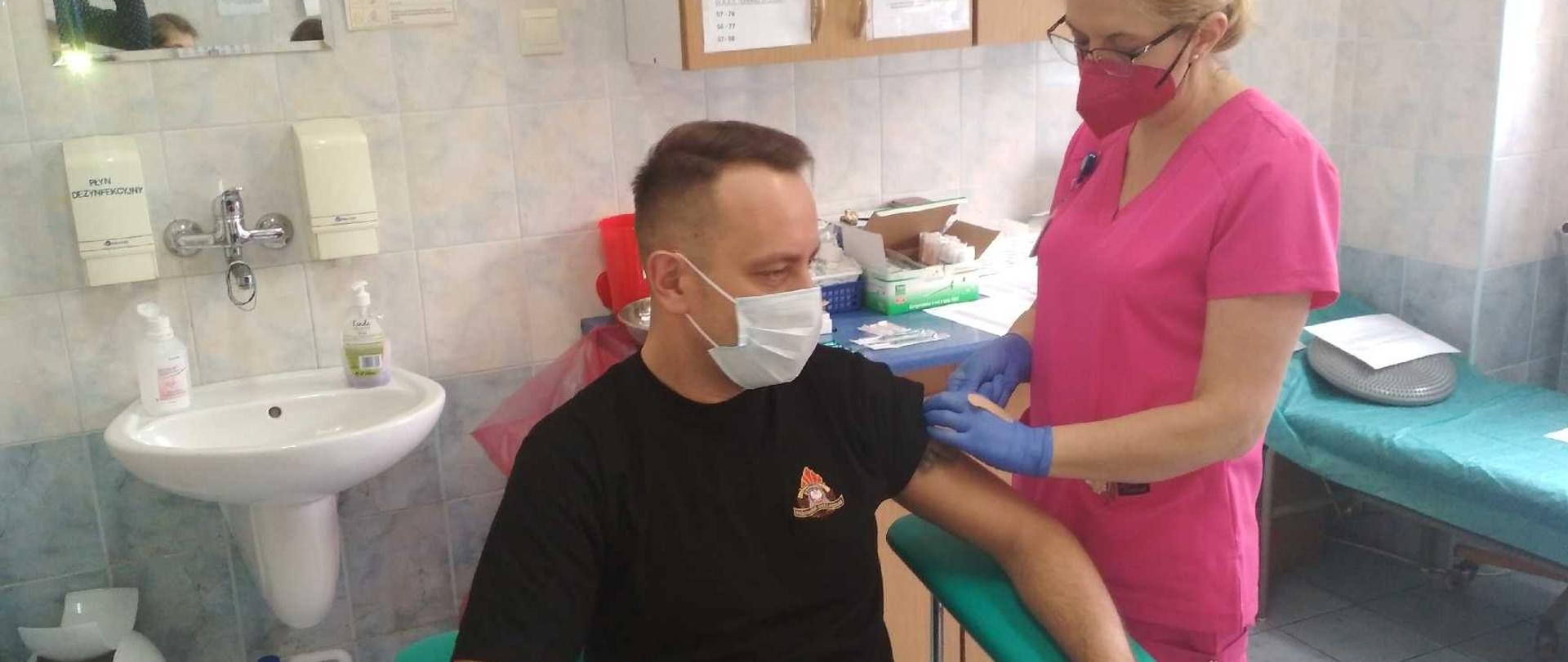 Zdjęcie przedstawia funkcjonariusza siedzącego na fotelu w gabinecie zabiegowym. Funkcjonariusz ubrany w koszulkę oraz spodnie od ubrania dowódczo – sztabowego. Ubranie jest w kolorze czarnym. Na twarzy posiada złożoną maseczkę. Obok niego po jego lewej stronie stoi pielęgniarka w ubraniu w kolorze różowym. Na twarzy ma również założoną maseczkę. Pielęgniarka szczepi funkcjonariusza szczepionką przeciwko covid-19. 