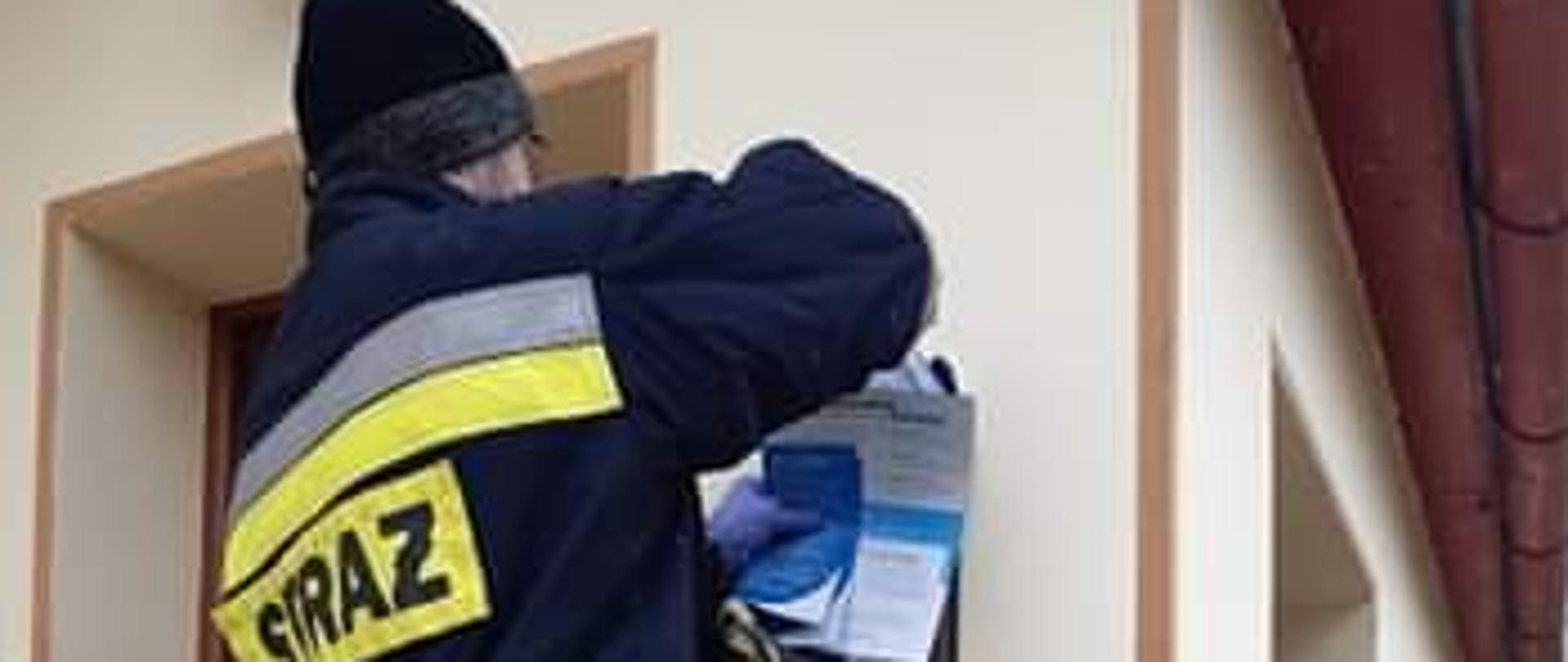 Strażak wrzuca ulotkę do skrzynki pocztowej zamontowanej do ściany budynku.