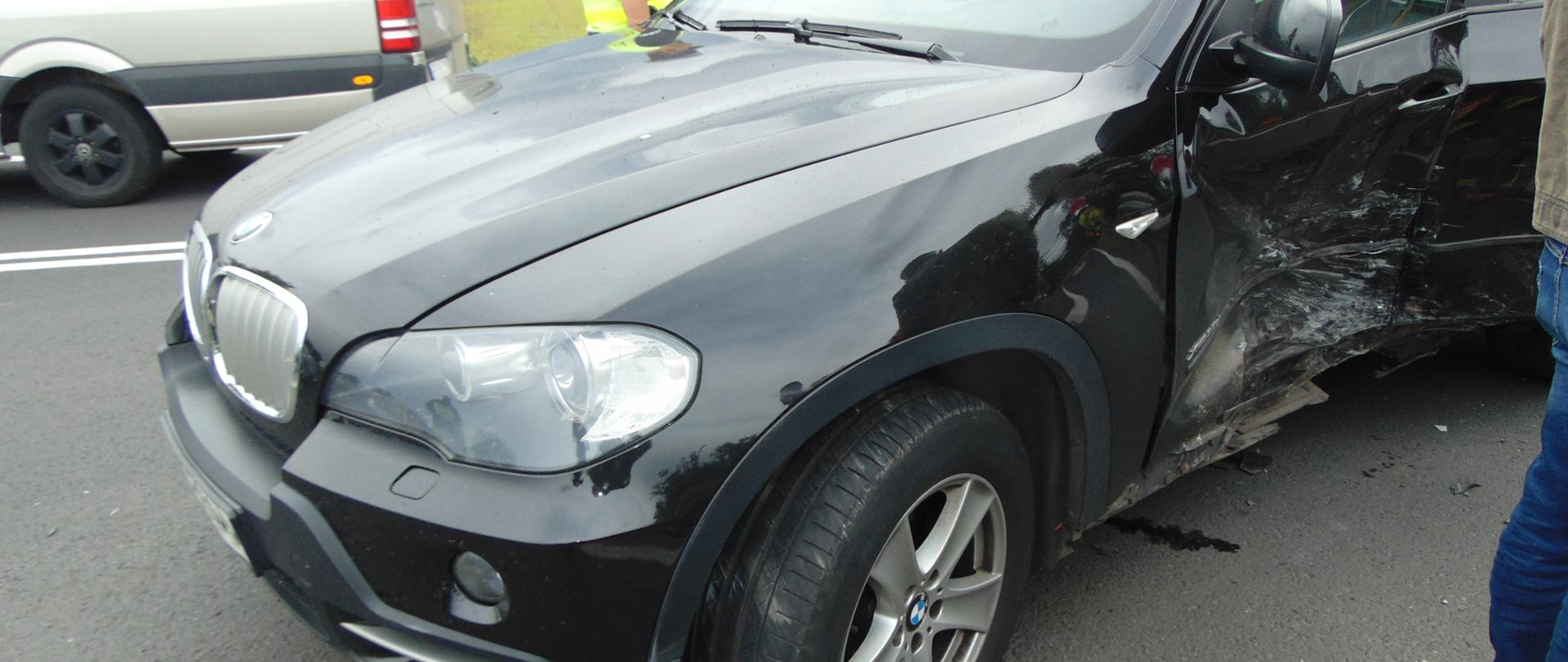 Samochód osobowy stoi na drodze z uszkodzonym na skutek zderzenia lewym bokiem pojazdu.