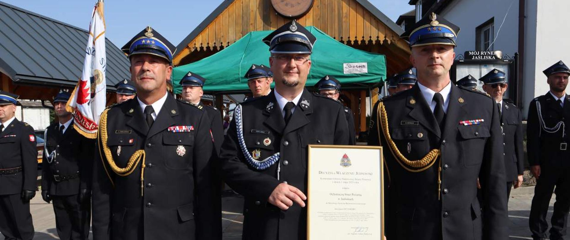 Na zdjęciu widoczny jest zastępca podkarpackiego Komendanta Wojewódzkiego PSP, Komendant Miejski PSP w Krośnie oraz prezes jednostki OSP Jaśliska prezentujący decyzję Komendanta Głównego PSP o włączeniu jednostki do Krajowego Systemu Ratowniczo – Gaśniczego. Na zdjęciu widoczne są również poczty sztandarowe w asyście strażaków OSP. Wszyscy strażacy występują w umundurowaniu wyjściowym. Impreza odbywa się na wolnym powietrzu, na rynku wyłożonym kostką brukową, w tle widać zabudowania miejscowości Jaśliska.