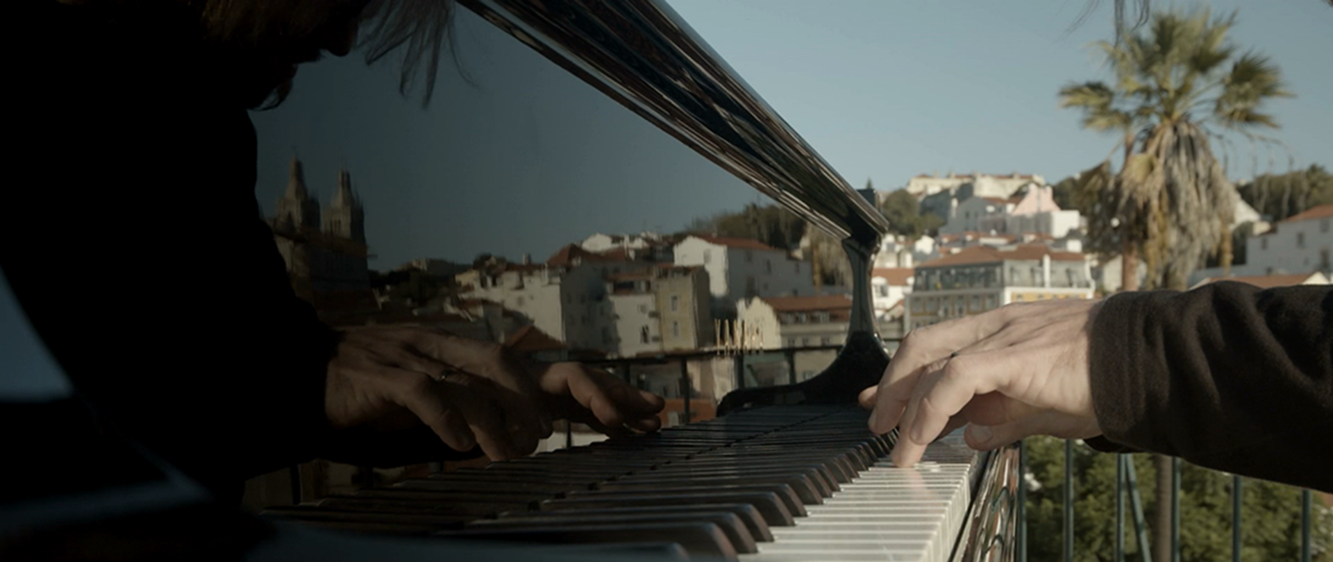 A imagem mostra as mãos de um pianista no teclado do piano. A vista de Lisboa reflete-se no instrumento.
