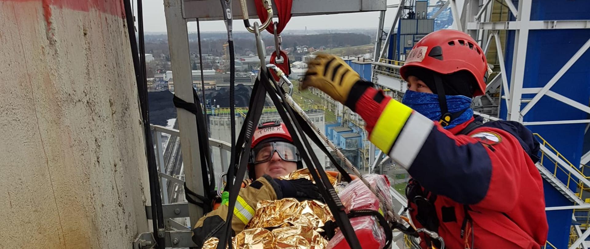 Na zdjęciu widzimy dwóch ratowników podczepionych na linach asekuracyjnych znajdujących się na znacznej wysokości.. Pierwszy ratownik znajdujący się z prawej strony kadru ubrany jest w specjalny kombinezon koloru czerwonego, na głowie czerwony kask. Asekuruje on drugiego strażaka, który pozoruje poszkodowanego. Drugi strażak znajduje się na desce ratowniczej koloru pomarańczowego w pozycji leżącej. Przykryty jest kocem termicznym koloru złotego.
Na głowie czerwony kask, okulary ochronne na oczach. Dalej w kadrze widzimy zabudowania przemysłowe.