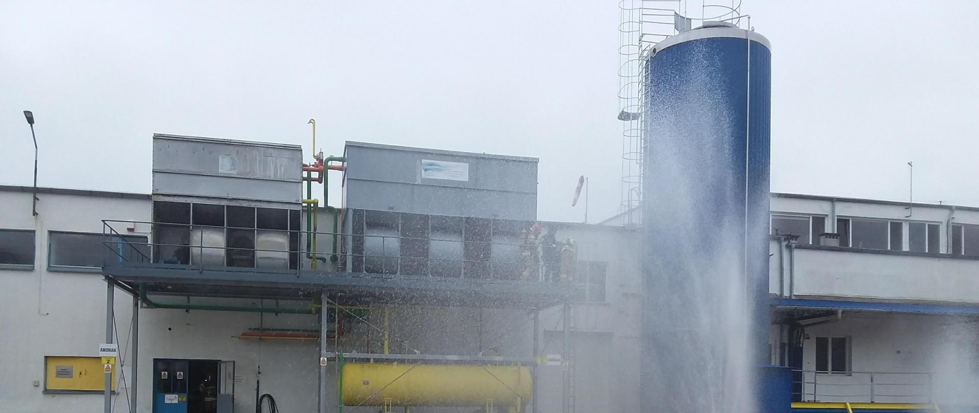 Plan zakładu produkcyjnego wyłożony betonowymi kostkami, w oddali widoczne budynki produkcyjne i instalacje technologiczne takie jak duże zbiorniki metalowe. Na placu przy pomocy armatury wodnej utworzona kurtyna wodna, za kurtyną widoczny zarys strażaków wchodzących po schodach. 