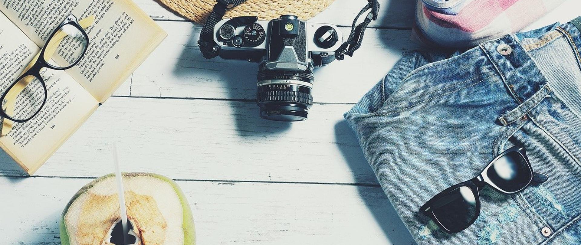 Grafika przedstawia książkę, okulary, kapelusz, letnią odzież, aparat i kokosa, położone na deskach