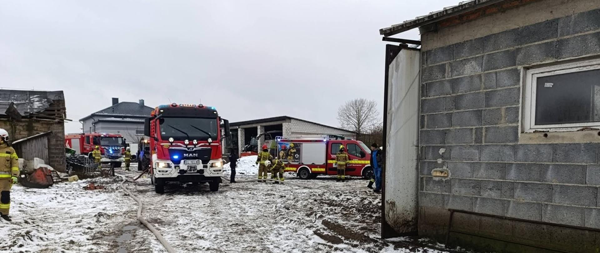 Na fotografii przedstawiono akcję służb ratowniczych prowadzoną na terenie gospodarstwa w zimowych warunkach. Centralnym punktem kadru jest wóz strażacki stojący na zaśnieżonym i błotnistym podłożu, z którego poprowadzony jest wąż gaśniczy w głąb terenu działań. W tle widoczne są kolejne pojazdy straży pożarnej oraz strażacy