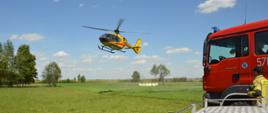 Śmigłowiec ratunkowy ląduje na łące w Dębnie Polskim. W tle drzewa, a na pierwszym planie fragmenty wozów strażackich i jeden ze strażaków uczestniczących w działaniach.