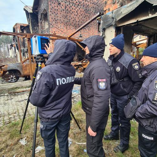 Instruktor i dwóch uczestników szkolenia przyglądają się funkcjonariuszce używającej skaner 3D do dokumentowania zniszczeń budynków