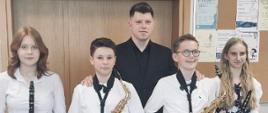 Na zdjęciu widzimy czworo uczniów, po środku stoi nauczyciel. Od lewej: Lena Kosielska (klarnet), Adrian Płochocki (saksofon), Mateusz Kurkowski (nauczyciel), Bartosz Surażyński (saksofon), Zuzanna Tarnacka (klarnet). Galowo ubrani uczniowie, trzymają w dłoniach instrumenty.