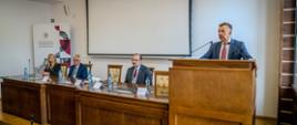 Minister Wieczorek stoi za mównicą i mówi do dwóch mikrofonów, obok niego za podłużnym drewnianym stołem siedzą trzy osoby.