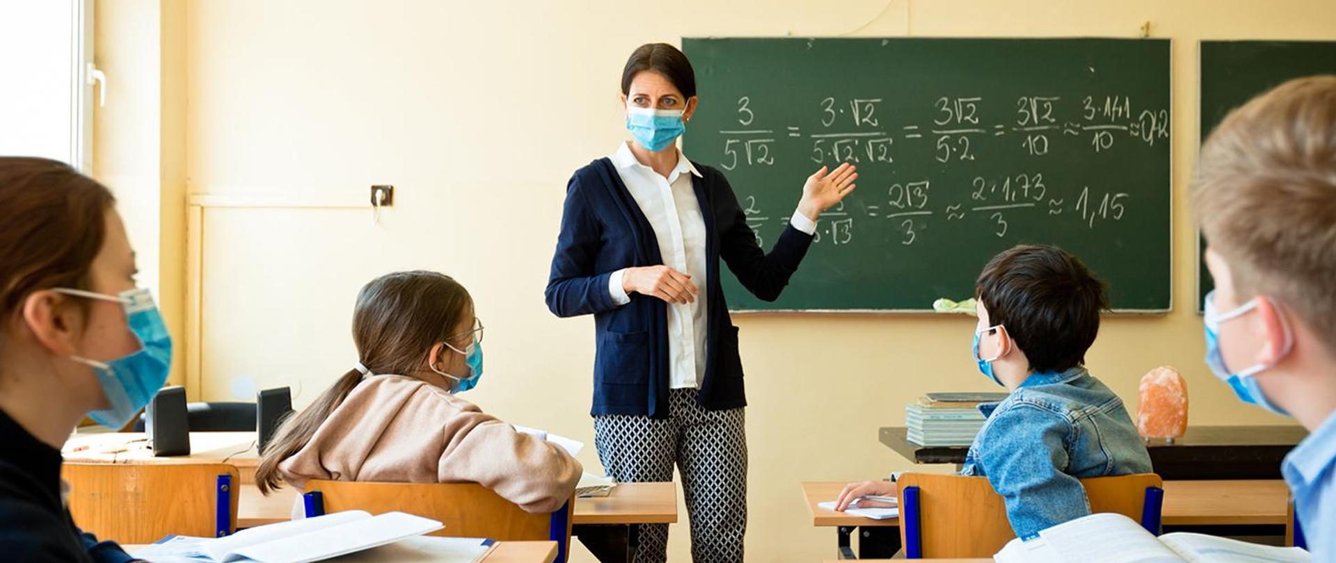 Nauczycielka matematyki tłumaczy w klasie czworgu uczniom ułamki. Uczniowie oraz nauczycielka w niebieskich maseczka