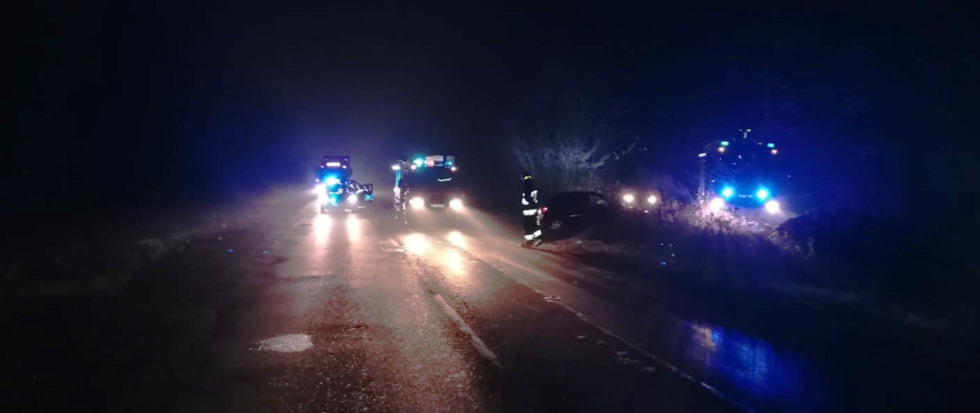 Zdjęcie przedstawia wozy strażackie z włączonymi światłami ostrzegawczymi podczas interwencji. W ciemnościach widać zarysy wozów strażackich i sylwetek strażaków.