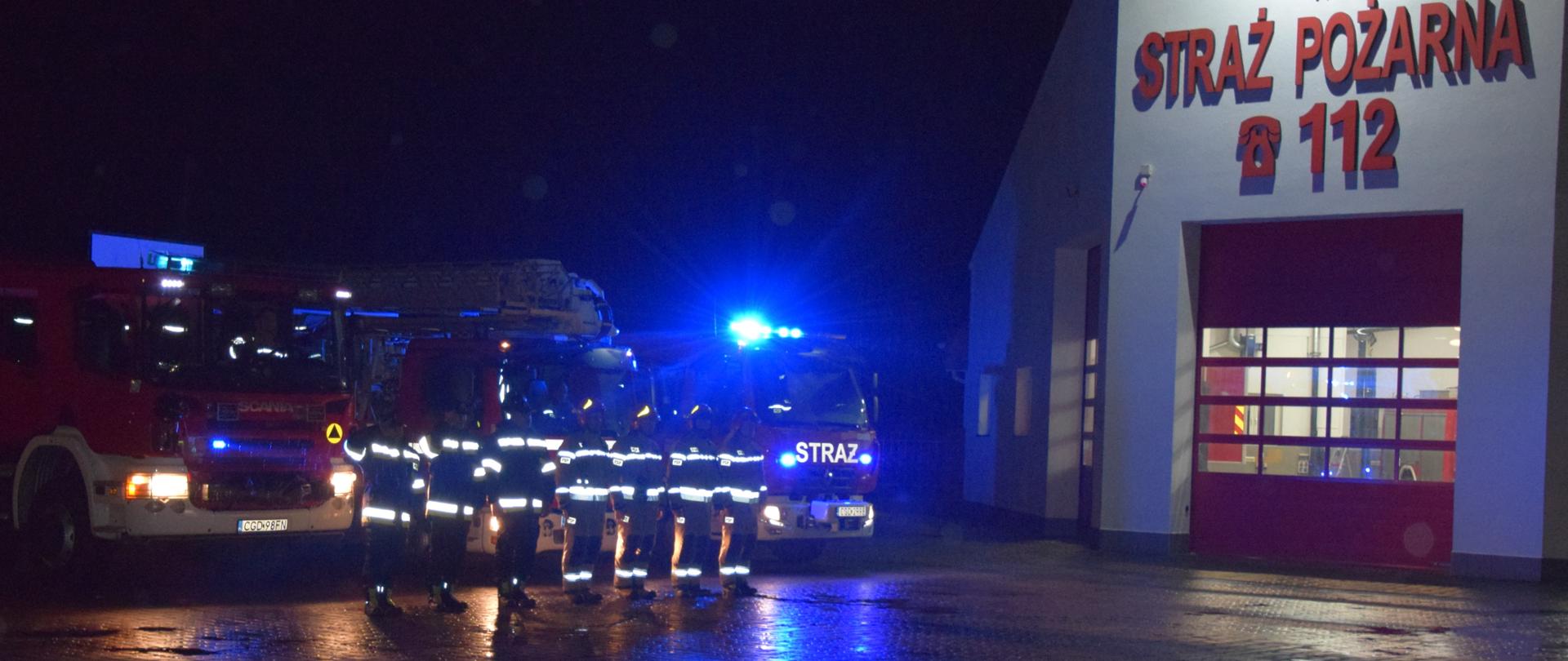 Na tle ustawionych wozów bojowych stoją strażacy w ubraniach specjalnych po prawej stronie widać fragment budynku strażnicy.