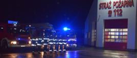 Na tle ustawionych wozów bojowych z włączonymi sygnałami świetlnymi stoją strażacy w ubraniach specjalnych po prawej stronie widać fragment budynku strażnicy.