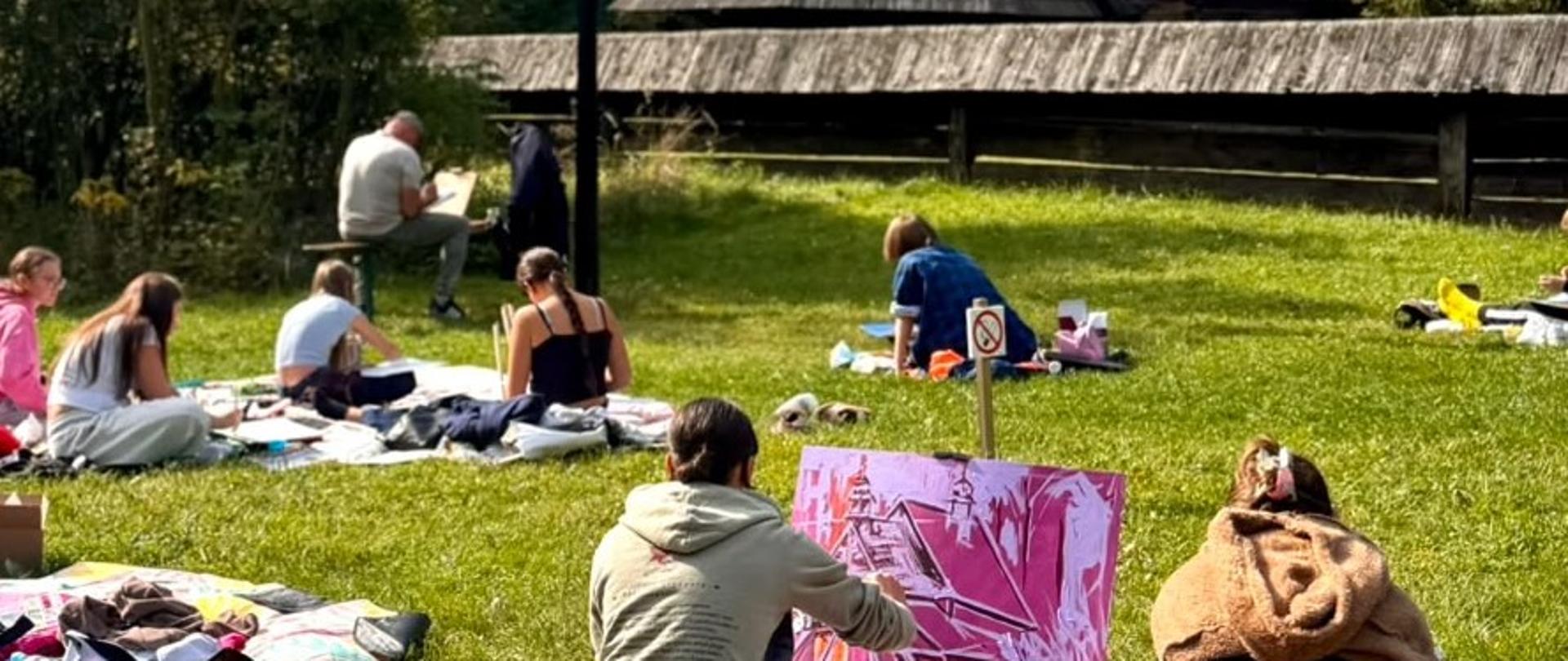 Na zdjęciu, w otoczeniu drzew, stoi drewniana, zabytkowa zabudowa sakralna z ciemnym, gontowym dachem i dwiema wieżyczkami zwieńczonymi krzyżami. Przed budynkiem na trawniku siedzą i leżą grupy osób, część z nich maluje lub rysuje. Na pierwszym planie kilka osób pracuje nad obrazami rozłożonymi na kocach. Pogodny dzień, błękitne niebo i jesienne drzewa tworzą spokojną, plenerową scenerię.