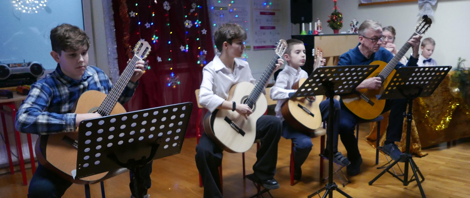 Na scenie zespół gitarowy, cztery osoby, dwaj chłopcy, dziewczyna i nauczyciel. Wszyscy grają siedząc na krzesłach, przed nimi pulpity nutowe.