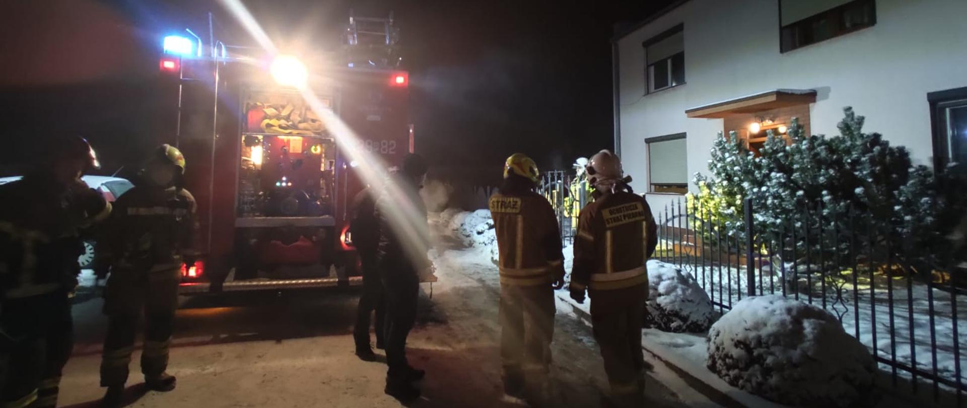 Na zdjęciu widać czerwony samochód strażacki przy którym stoją strażacy. Jest pora nocna, samochód strażacki oświetla teren akcji. Po prawej stronie za ogrodzeniem widać jasny budynek wielorodzinny