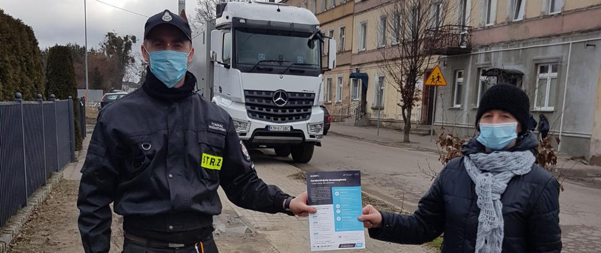 Zdjęcie przedstawia strażaka OSP przekazującego ulotkę informacyjną o szczepieniach przeciwko COVID-19. W tle budynki mieszkalne i samochód ciężarowy.