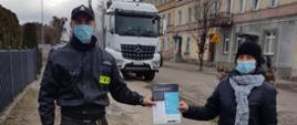 Zdjęcie przedstawia strażaka OSP przekazującego ulotkę informacyjną o szczepieniach przeciwko COVID-19. W tle budynki mieszkalne i samochód ciężarowy.