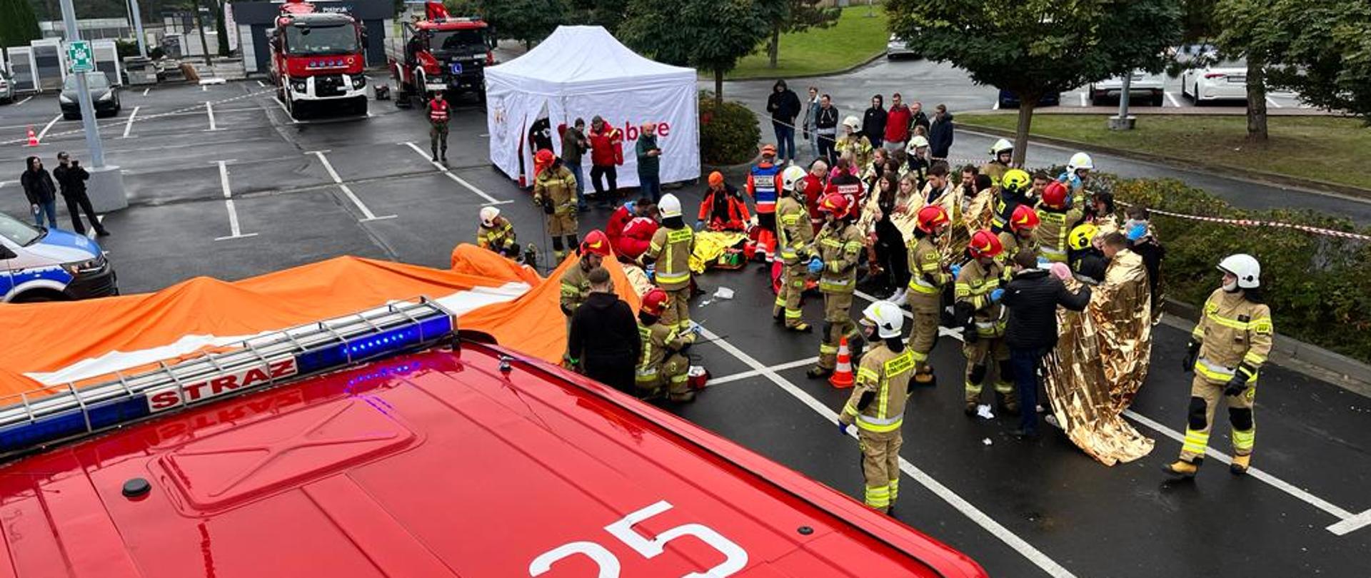 Dach samochodu pożarniczego. W tle służby ratownicze zaopatrują poszkodowanych.