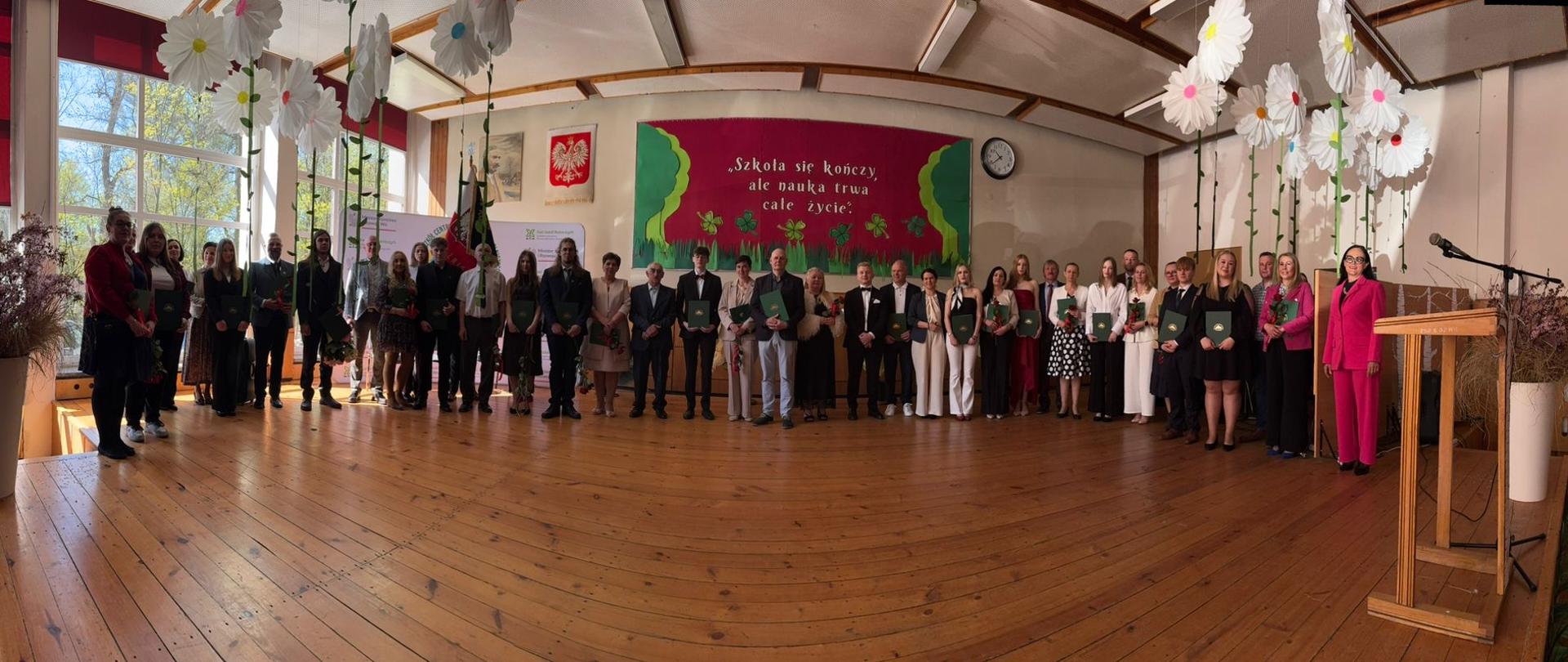 Panoramiczne zdjęcie grupowe na szkolnej scenie. W długim rzędzie stoją uczniowie w strojach galowych trzymający dyplomy i kwiaty, ich rodzice, nauczycielki w eleganckich strojach oraz dyrekcja. Scena ozdobiona jest wiszącymi, wielkimi białymi kwiatami z papieru. Na ścianie w tle widnieje czerwona tablica z napisem: „Szkoła się kończy, ale nauka trwa całe życie” oraz godło Polski. Po prawej stronie widać drewnianą mównicę i mikrofon.