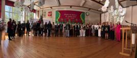 Panoramiczne zdjęcie grupowe na drewnianej scenie auli. W długim rzędzie stoją wyróżnieni uczniowie w strojach galowych, trzymając świadectwa i kwiaty, a obok nich dyrekcja szkoły, nauczyciele oraz rodzice. Aula jest bogato udekorowana dużymi, białymi papierowymi kwiatami zwisającymi z sufitu. W tle na ścianie widać bordową tablicę z hasłem „Szkoła się kończy, ale nauka trwa całe życie” oraz godło Polski. Z boku stoi drewniana mównica.