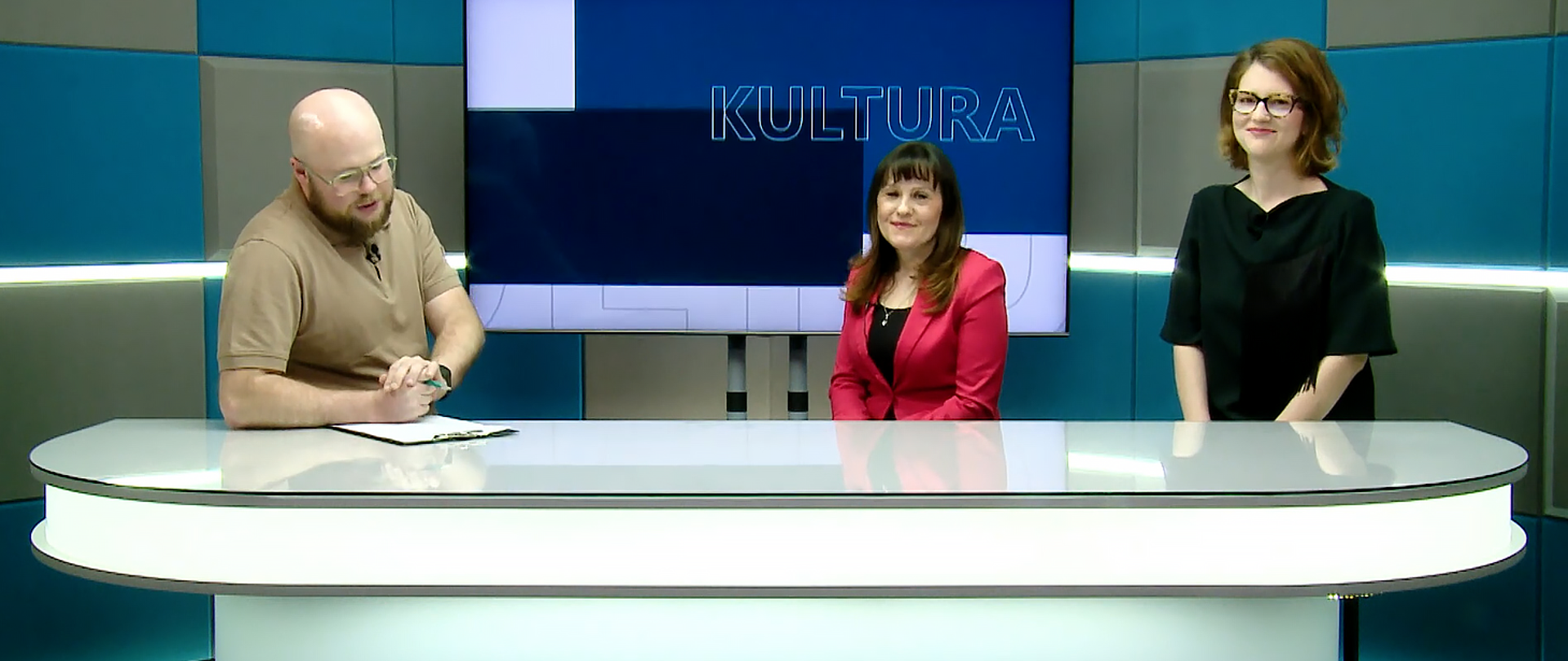 Studio telewizyjne. W tle na niebiesko-szarej ścianie wisi telewizor a na nim napis "kultura", z przodu przy białym, podłużnym stole siedzą trzy osoby, z lewej mężczyzna patrzący na kartkę leżącą na stole, z prawej strony dwie kobiety patrzące w kierunku widza.