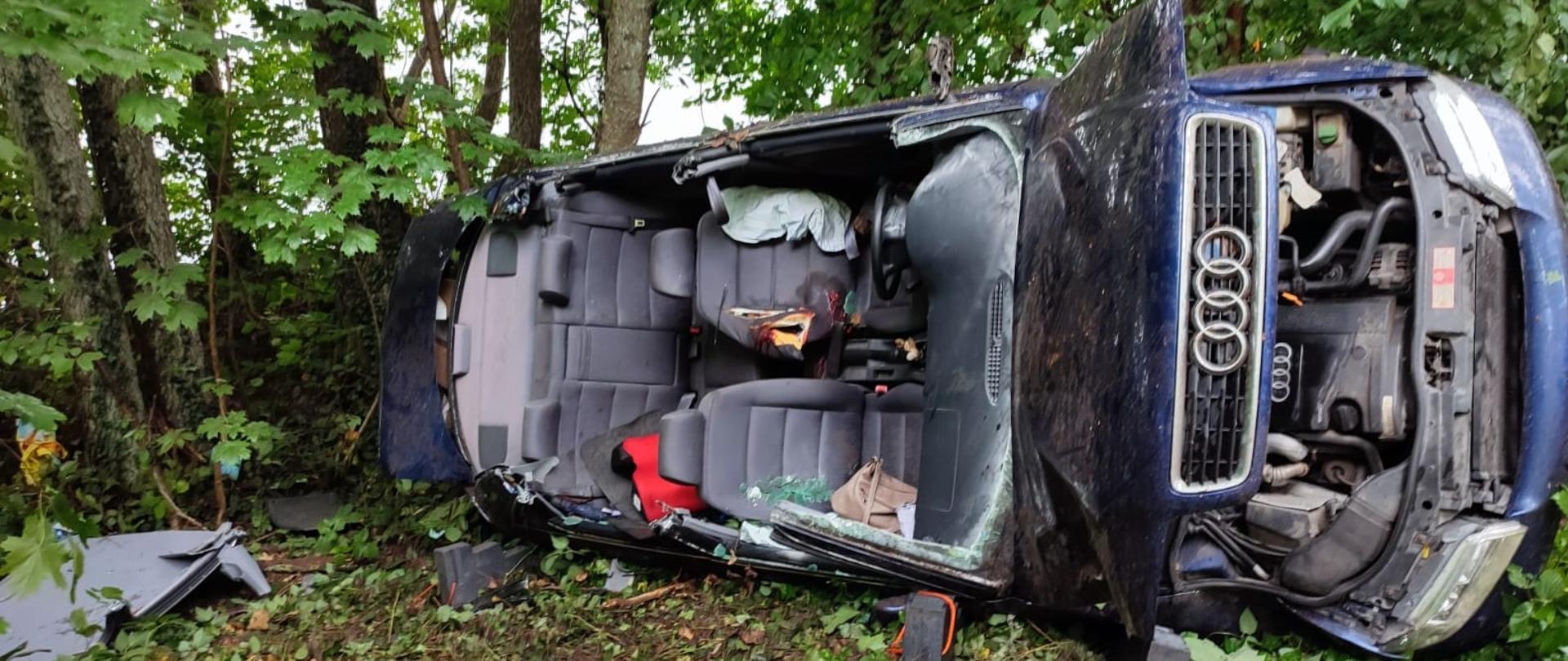 Samochód osobowy marki audi, ciemnego koloru leży na boku. Ma wycięty dach w środku nikogo nie ma.