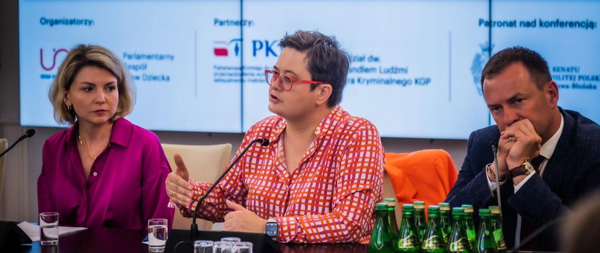 wiceminister Lubnauer przemawia w czasie spotkania na siedząco przy stole konferencyjnym. z lewej strony siedzi kobieta w różowej sukience z prawej mężczyzna w garniturze. 