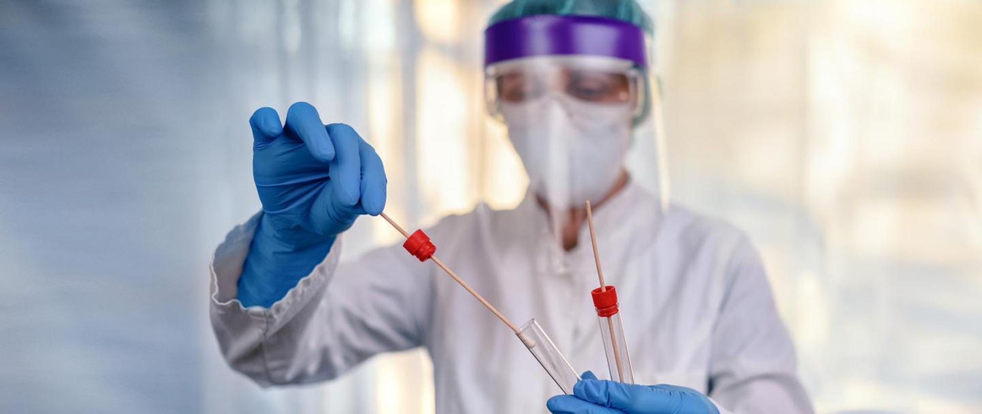 Na jasnym tle kobieta w białym fartuchu, maseczce i przyłbicy, w niebieskich rękawiczkach jednorazowych. W rękach trzyma dwie probówki do wymazu