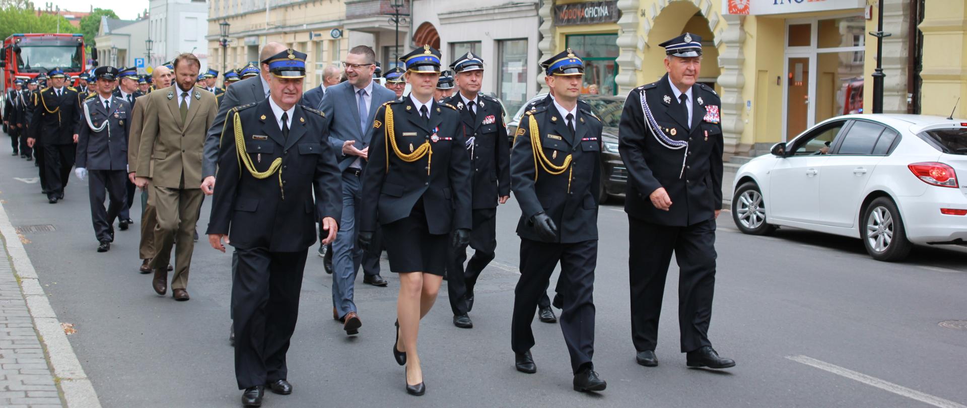 grupa strażaków idzie ulicą, przemarsz, w tle kamienice i samochód strażacki