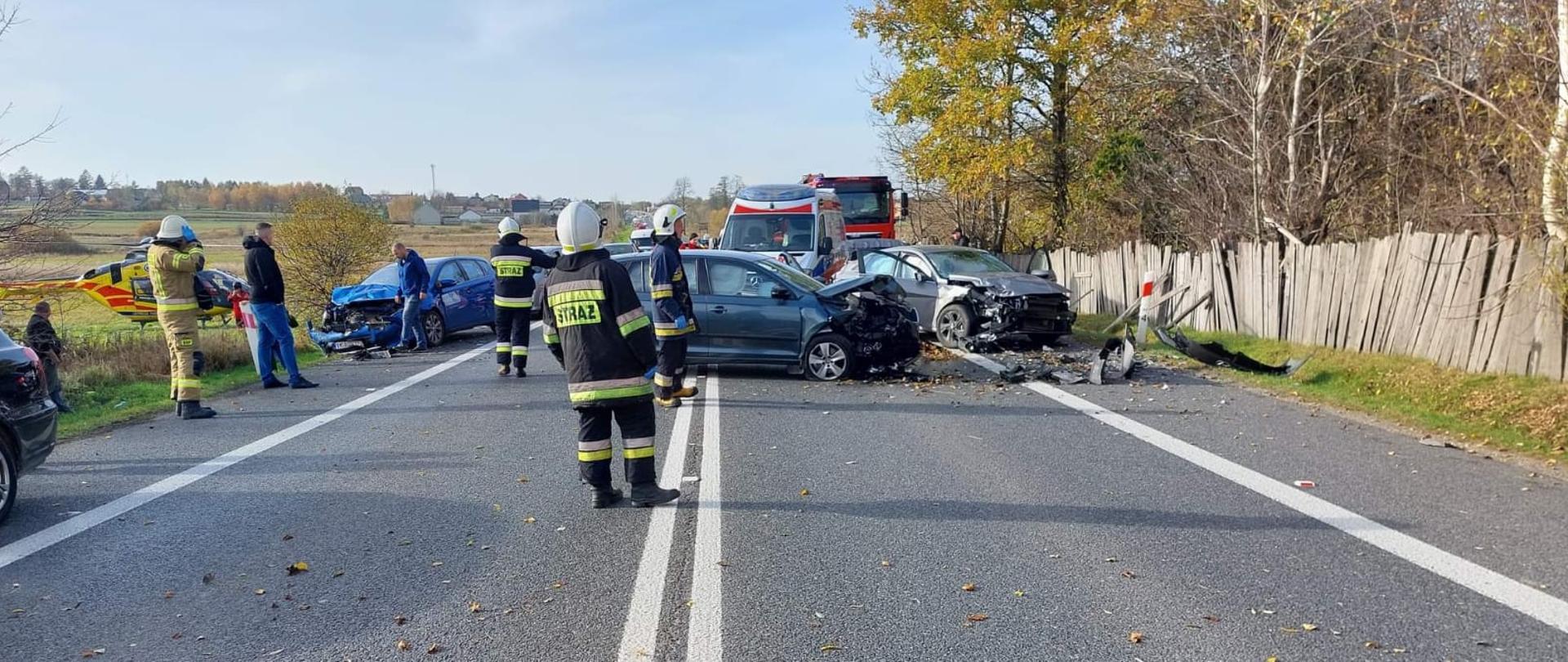 Zdjęcie przedstawia krajową 74 na której doszło do wypadku komunikacyjnego. Głównym elementem zdjęcia są trzy uszkodzone auta. Po lewej stronie, na łące stoi śmigłowiec LPR-u. Przed autami przemieszczają się strażacy i osoby postronne, a za uszkodzonymi pojazdami widać ambulans i wóz strażacki.