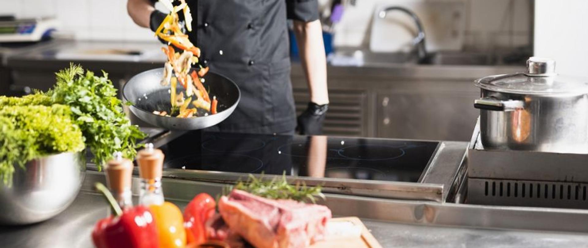 pomieszczenie kuchni w zakładzie gastronomicznym. na pierwszym planie leży miska na której znajduje się mięso, papryki i przyprawy. na dalszym planie kucharz smaży warzywa na patelni