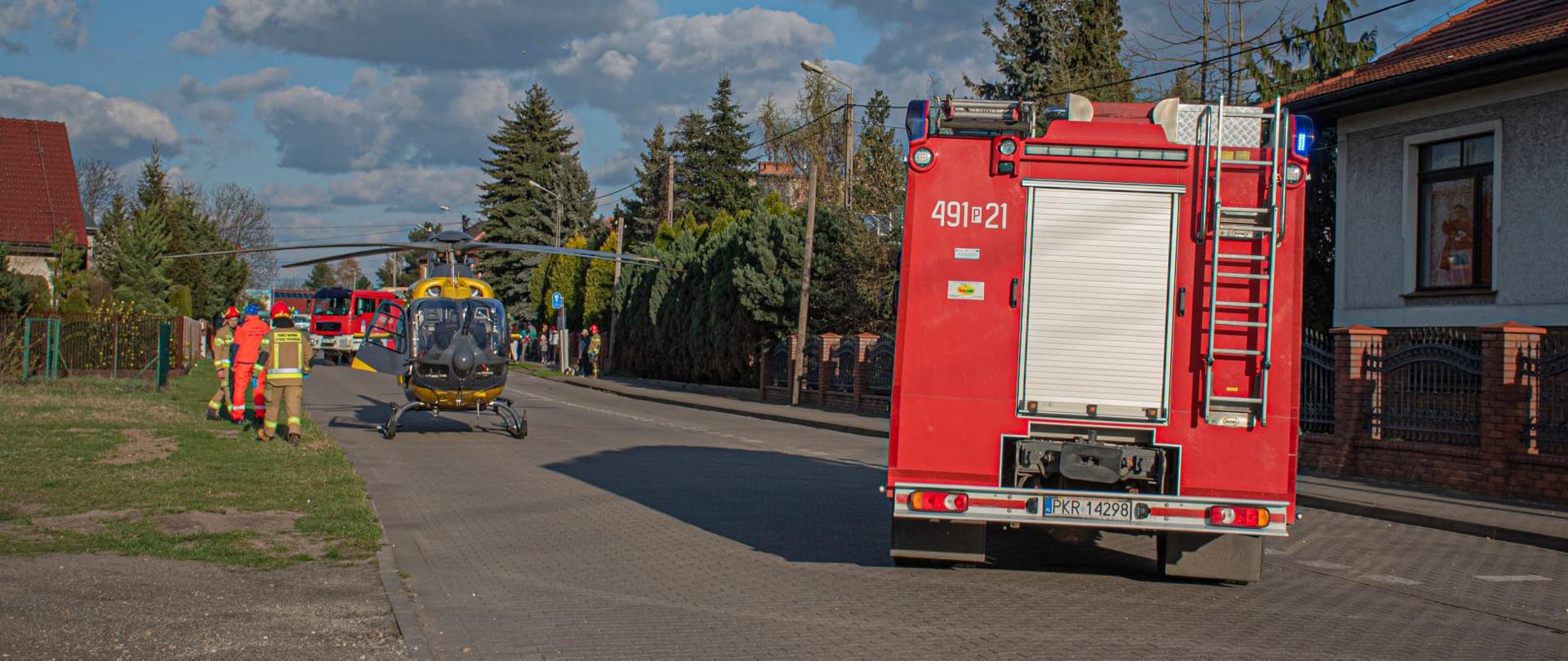 Na drodze wewnętrznej stoi śmigłowiec LPR oraz dwa wozy straży pożarnej, które zabezpieczają lądowisko. Obok śmigłowca stoją strażacy wraz z ratownikiem medycznym. W oddali widać grupę ludzi obserwującą całe zdarzenie. 