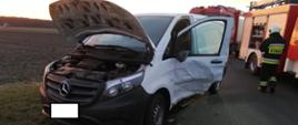 Zdjęcie przedstawia samochód dostawczy koloru białego z uszkodzonym lewym bokiem na skutek zdarzenia drogowego w tle samochody straży pożarnej.