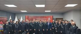 Na zdjęciu znajduje się duża grupa strażaków w galowych mundurach, ustawiona w kilku rzędach w sali konferencyjnej. W tle widoczne są czerwone ścianki z emblematami Państwowej Straży Pożarnej oraz biało-czerwone flagi ustawione na stojakach. Pośrodku grupy stoją również osoby w garniturach. Pomieszczenie ma jasne, sufitowe oświetlenie, a przed grupą widoczne są stoły i krzesła. Scena ma charakter oficjalny i uroczysty.