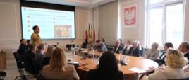 Szeroki kadr jasnej, przestronnej sali konferencyjnej. Wokół dużego, owalnego, drewnianego stołu siedzi kilkanaście osób – dyrektorzy i przedstawiciele placówek oświatowych w strojach wizytowych oraz funkcjonariusze służb. U szczytu stołu stoi oficer Państwowej Straży Pożarnej w piaskowym mundurze służbowym. Prowadzi prelekcję na tle dużego ekranu projekcyjnego, na którym wyświetlany jest slajd prezentacji dotyczący wyznaczania miejsc schronienia w budynkach. Na ścianie po prawej stronie wisi godło Polski w ramie, obok stoją flagi państwowe i samorządowe. Na stole leżą materiały szkoleniowe, notesy oraz karafki z wodą. Uczestnicy spotkania uważnie słuchają wykładu i patrzą w stronę prelegenta.