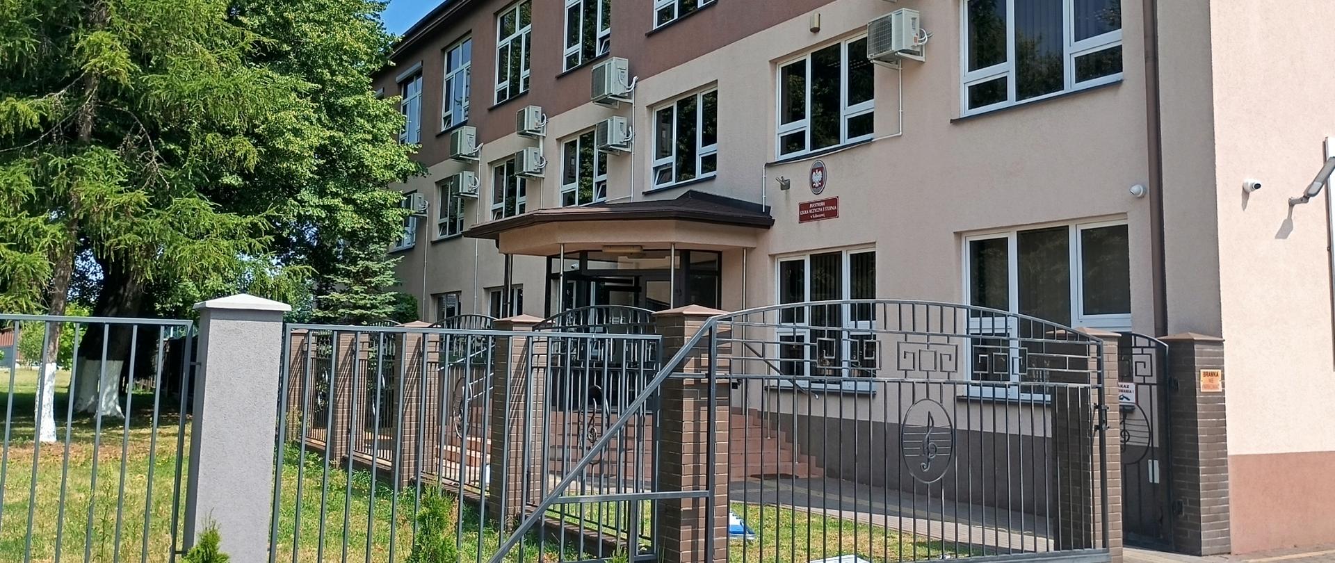 Budynek szkoły w kolorze kremowo brązowym z czarnym ogrodzeniem.