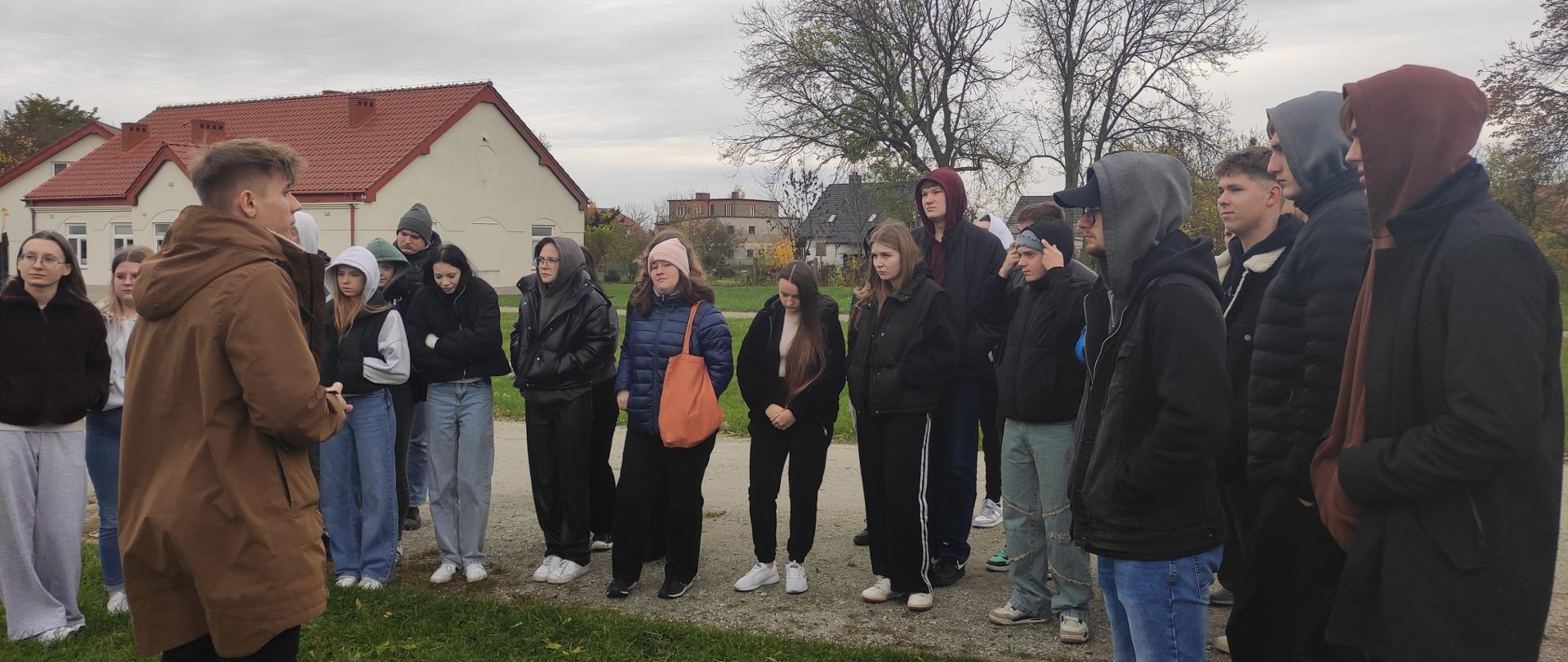 Grupa kilkudziesięciu uczniów stoi na zewnątrz w chłodny dzień, słuchając osoby prowadzącej zajęcia. Młodzież ubrana jest w kurtki i bluzy z kapturem, a w tle widać budynki z czerwonymi dachami i bezlistne drzewa.
