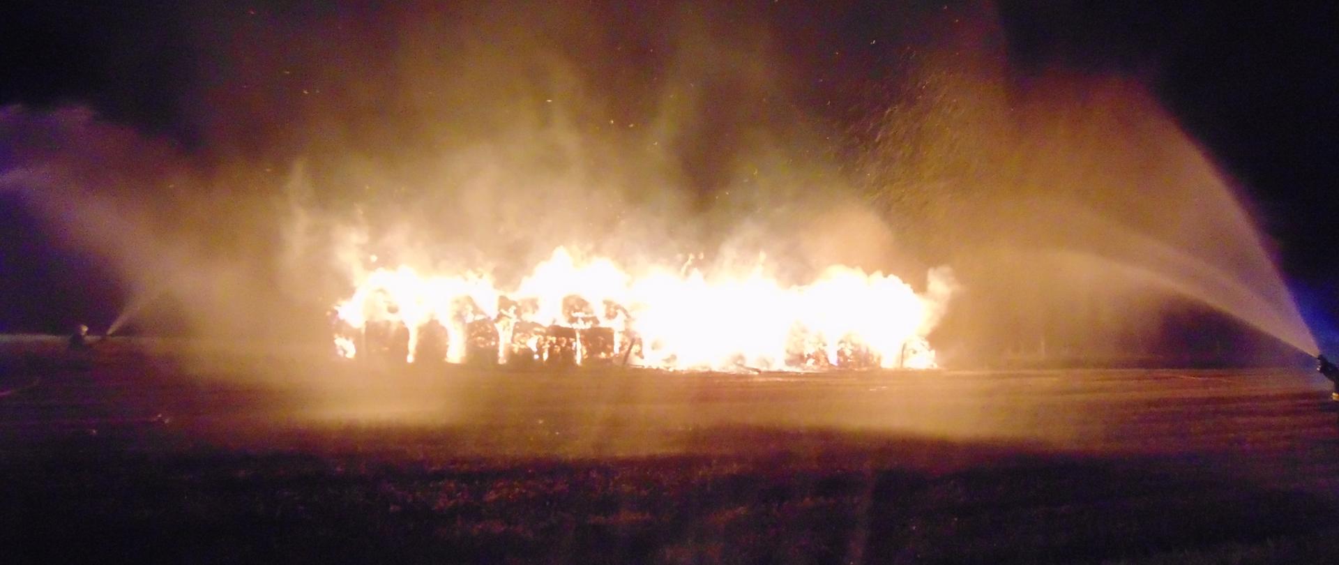 Pali się stóg balotów słomy. z dwóch stron strażacy podają dwa prądy wody na pożar.