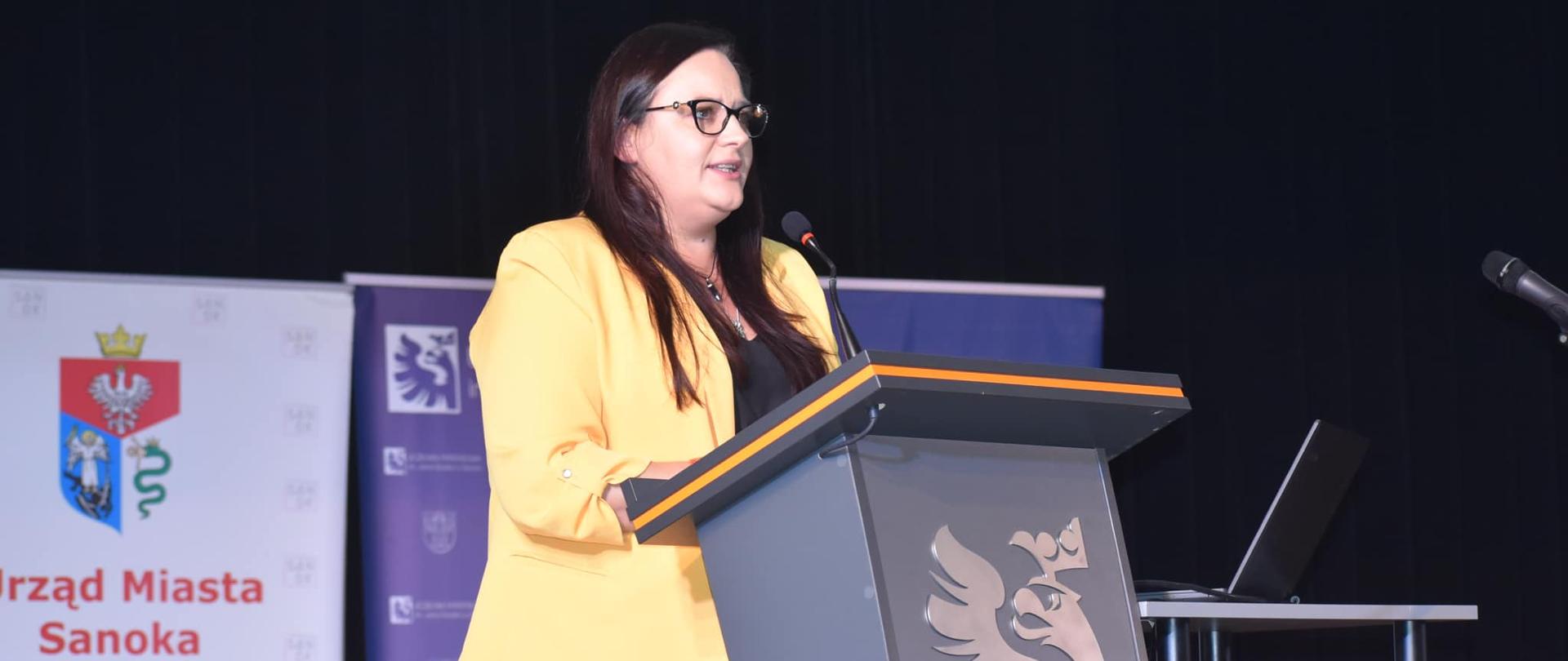 Wiceminister Małgorzata Jarosińska-Jedynak stoi w mównicy z mikrofonem. Za nią banery.